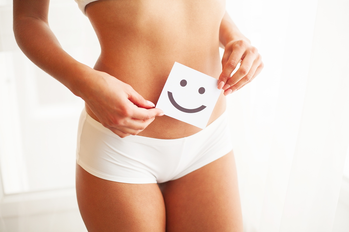 Femeie în lenjerie albă ținând un card cu zâmbet pe abdomen — confort și echilibru intim